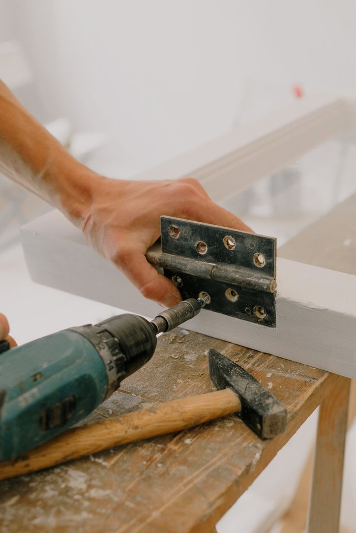 Close-up of an anonymous craftsman fixing a metal hinge to a wooden window frame using a contemporary screwdriver, set on a wooden board amid renovation activities